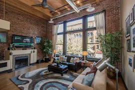 Staged living room with large picture windows, brick accent walls, exposed ceilings and vents, ceiling fan, fireplace