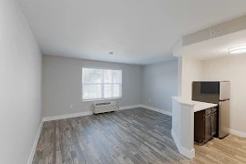 Living room and bedroom area with light walls, white trim, wood-inspired flooring, window with blinds, and access to kitchen