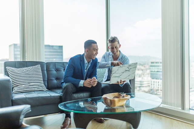 Engaged customers - two men pointing and laughing at laptop as they sit on couch in high-rise building.