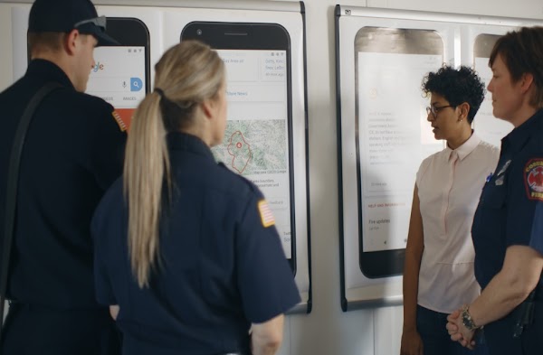 A Googler and three first responders stand in front of posters displaying examples of how a wildfire boundary map appears on Google Search