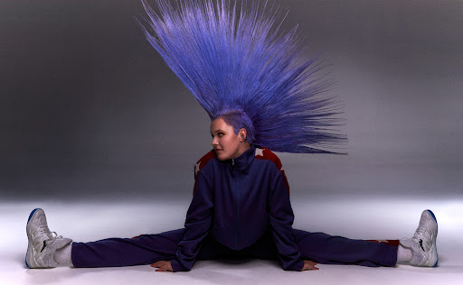 Jojo Siwa with a large blue mohawk in a matching blue track suit doing the splits on the ground.