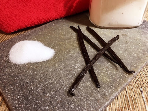 Sugar and a few vanilla beans on a grey cutting board.