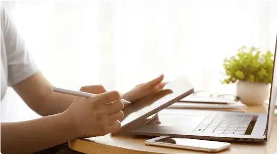 O proprietário de uma empresa trabalhando em casa em um tablet. Também estão sobre a mesa, à frente dele, um laptop aberto e um smartphone.