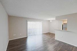 Living room with wood plank floors, sliding glass patio door with blinds, and light gray walls, next to kitchen