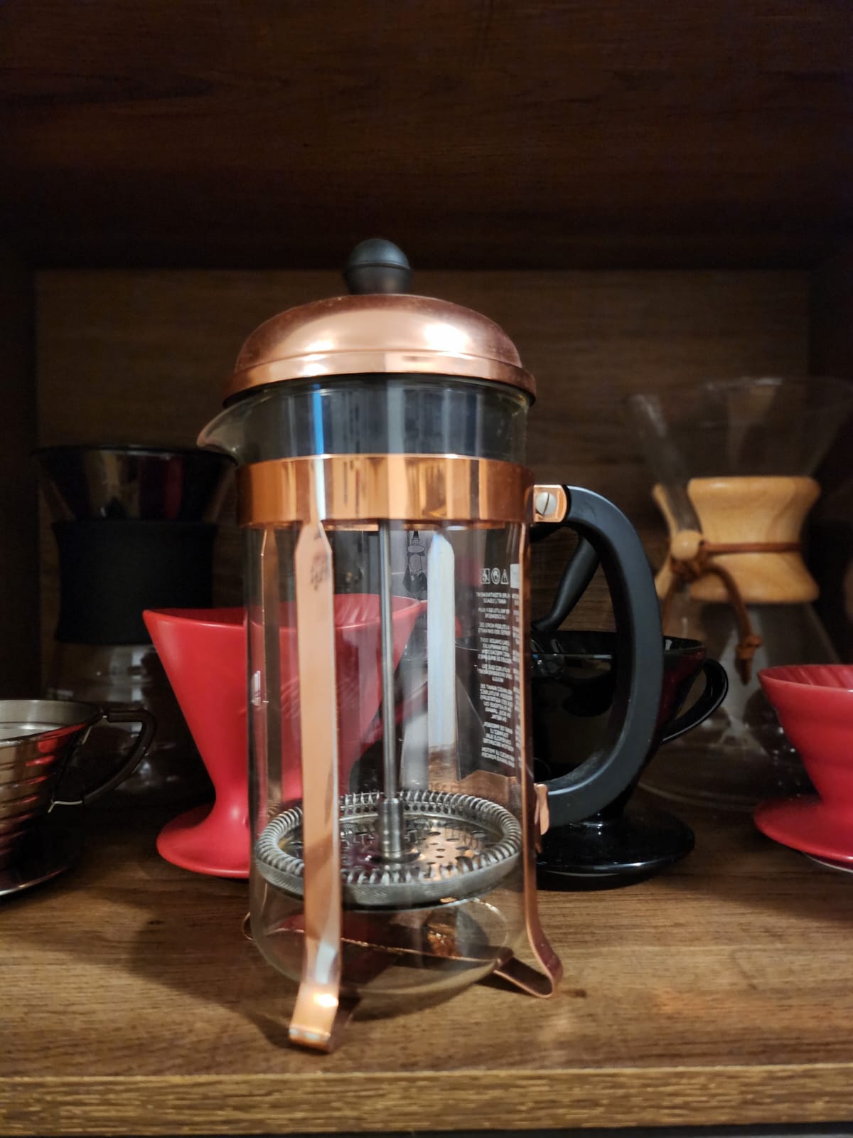 Foto de French Press, com corpo feito em vidro transparente, alça em plástico preto e detalhes do corpo e tampa em metal da cor rosê.
Imagem com filtro French Press em destaque, com fundo de madeira. Outros métodos diversos de café ao fundo, desfocados.