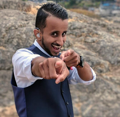 Narada grins playfully at the camera, pointing his fingers at the person shooting the picture. He is a young man wearing a collared white shirt, a black vest, and a hearing aid. He stands in front of a rocky landscape outside.