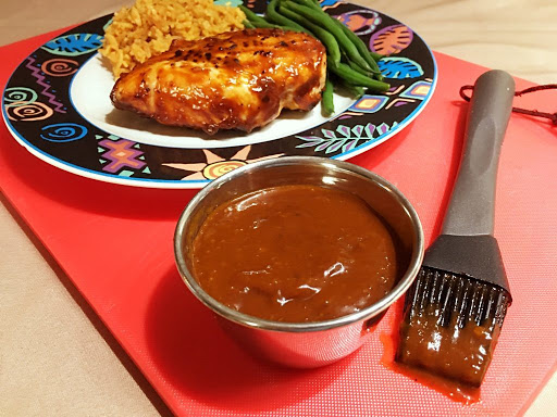 A bowl filled with sauce next to a brush with chicken breast, rice and green beans on a plate in the background.