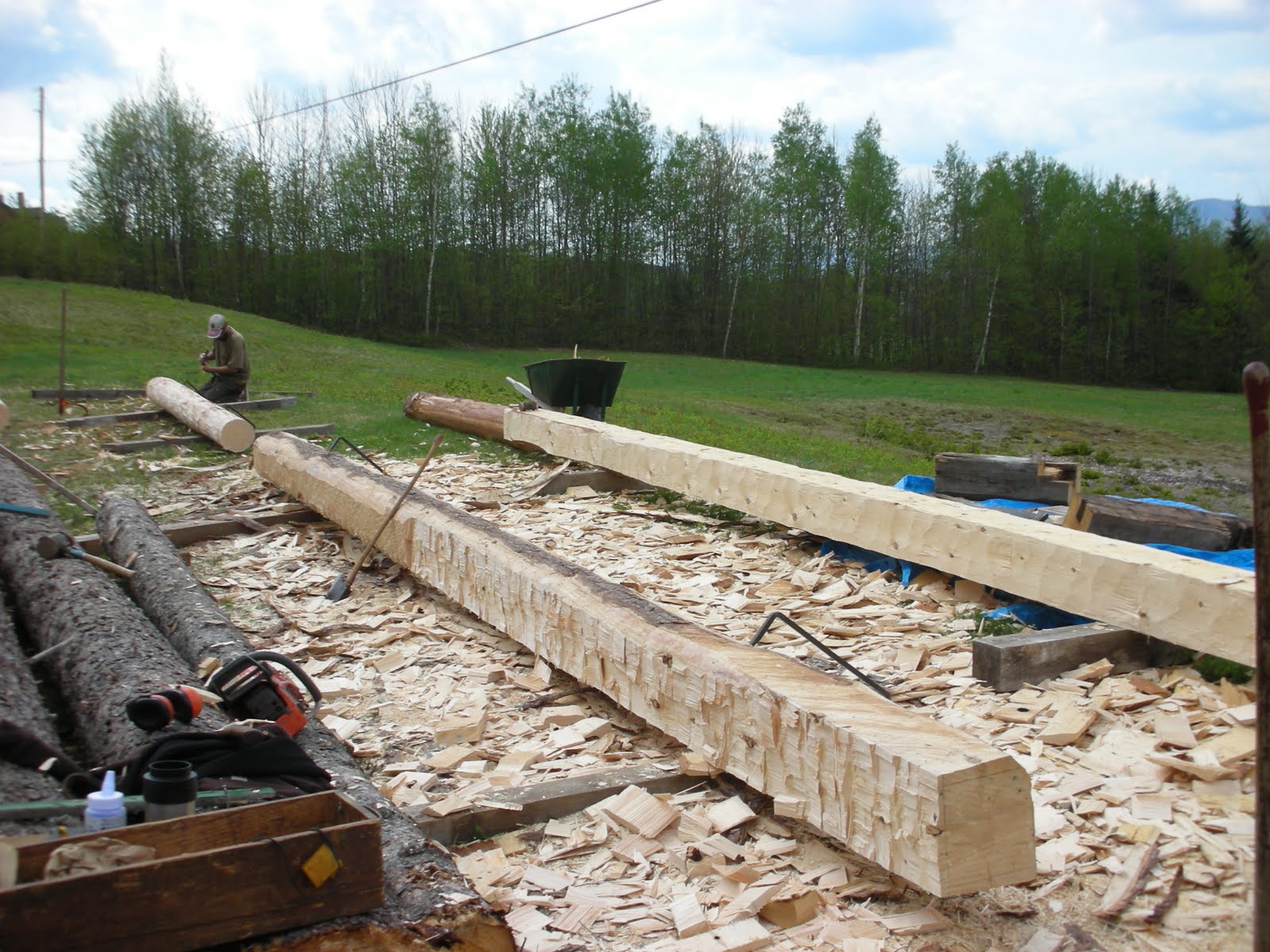 The finished sill timber is in the background with a new sill being roughed out in the foreground.  Michael begins laying out a post timber from a log.
