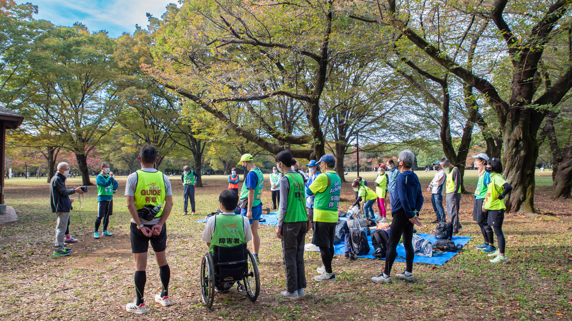 秋晴れの日の代々木公園に集まる約20人のアキレス・インターナショナル・ジャパンのみなさん。マイクで話す男性に耳を傾けている。