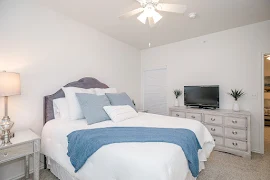 Model bedroom with plush carpet, white walls, large bed with white linens, and ceiling fan