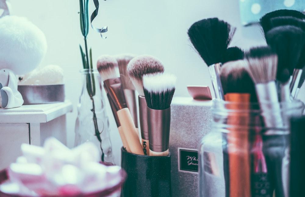 A sparkly cup of makeup brushes surrounded my a vase with a plant, and a jar of more makeup brushes