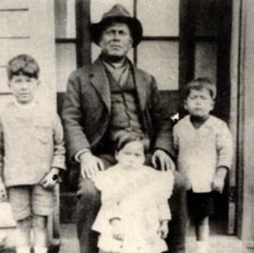 [Fotografía] en blanco y negro de tres niños pequeños, Gonzalo Rojas y dos de sus hermanos, alrededor de un adulto con sombrero,
el padre, en un porche mirando al frente.