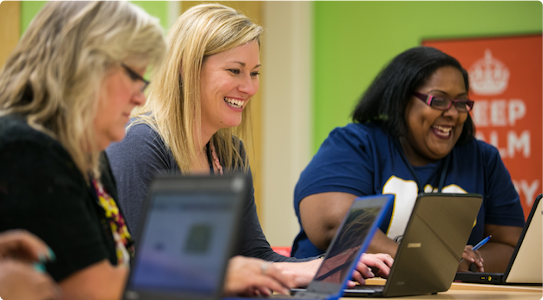 Three adult women sit at a long table with their laptops, laughing.