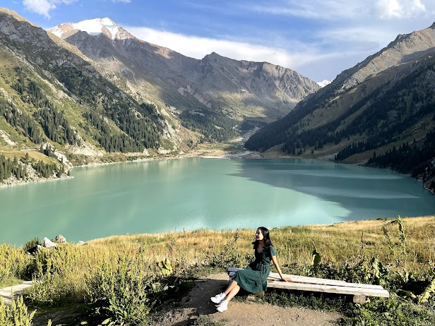 Photo of Aliya Rysbek sitting on a wooden bench in front of a lake surrounded by mountains. There is snow on the peak of the tallest.