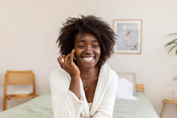 ãoDaImagem: mulher preta tem cabelos cacheados, sorri enquanto passa produto no rosto; ela veste um roupão branco e ao fundo é possível ver mobília de um quarto. Foto: Freepik.