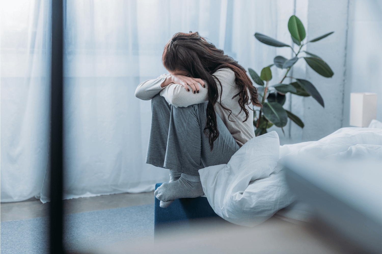 Woman sitting on edge of bed clutching her knees and resting her head on her arms