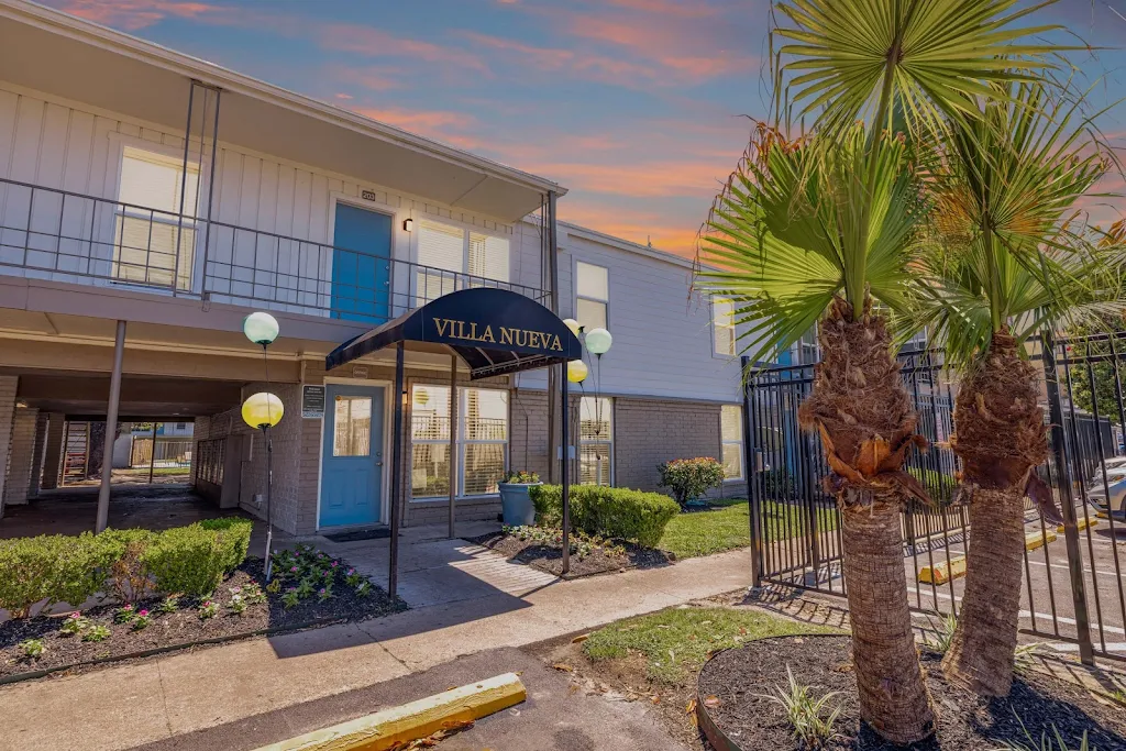 Apartment building entrance with "Villa Nueva" sign, lit pathway, palm trees, and colorful sky at dusk.