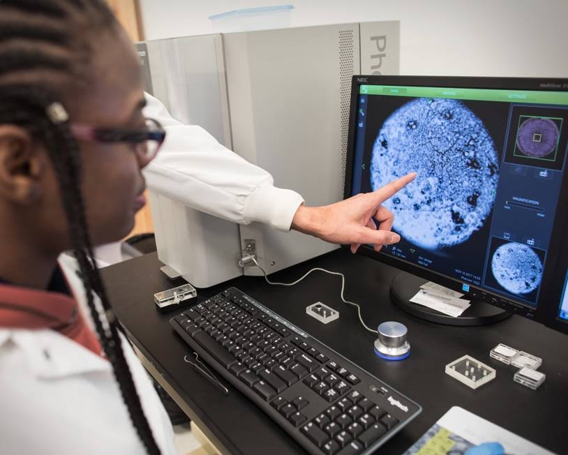 Dito puntato sullo schermo di un computer con una donna che guarda da vicino. L'ambientazione sembra un laboratorio di scienze durante l'osservazione dell'ingrandimento di una massa circolare.