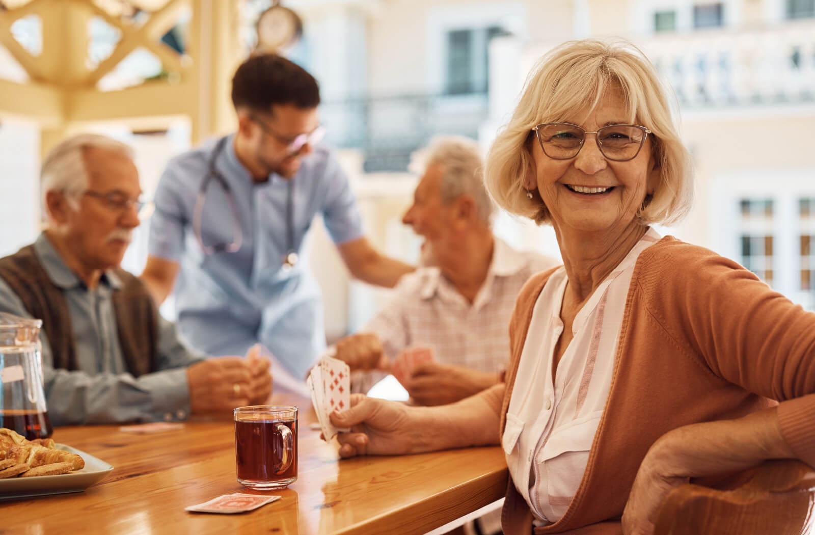 A senior woman sits at a table, holding cards next to a cup of tea and smiling. In the background, fellow seniors engage in conversation while a nurse helps out.
