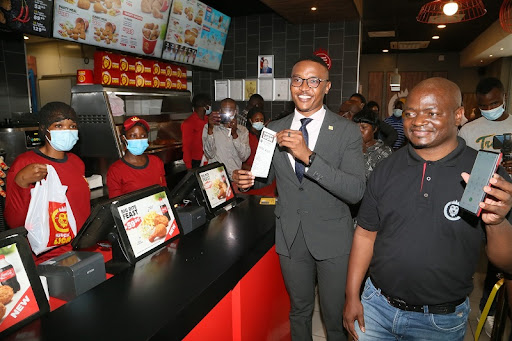 MTN Zambia Mobile Money Managing Director Komba Malukutila and Hungry Lion Country Manager Busu Mpepo showing how it is done to buy using MTN money shortly after the official launch of the partnership with Hungry Lion and MTN mobile money at Pinnacle Mall in Lusaka yesterday. Thursday, November 25, 2021.  Picture by ROYD SIBAJENE/ZANIS.