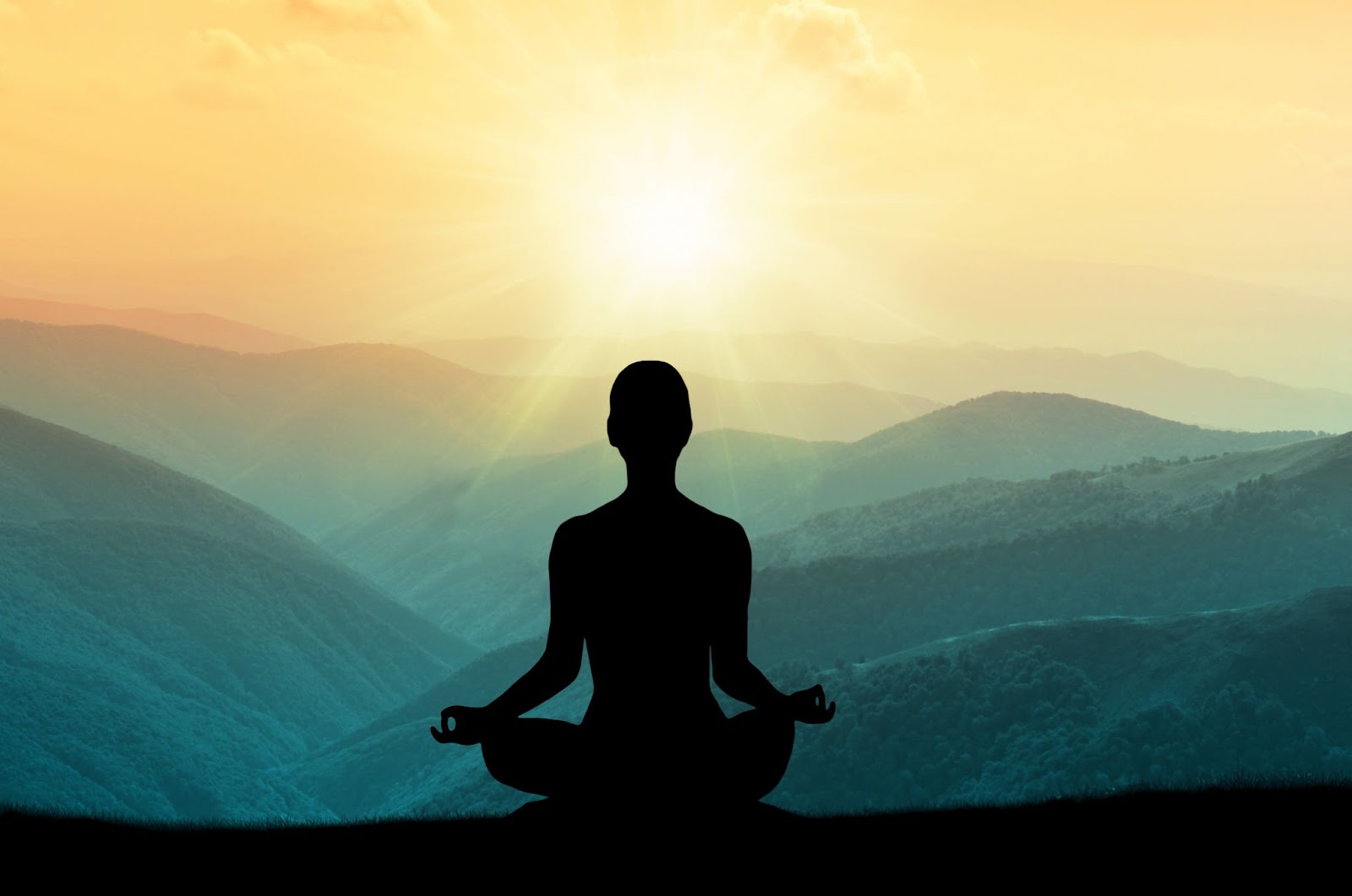 A silhouette of a person meditating overlooking hills with the sun shining. 