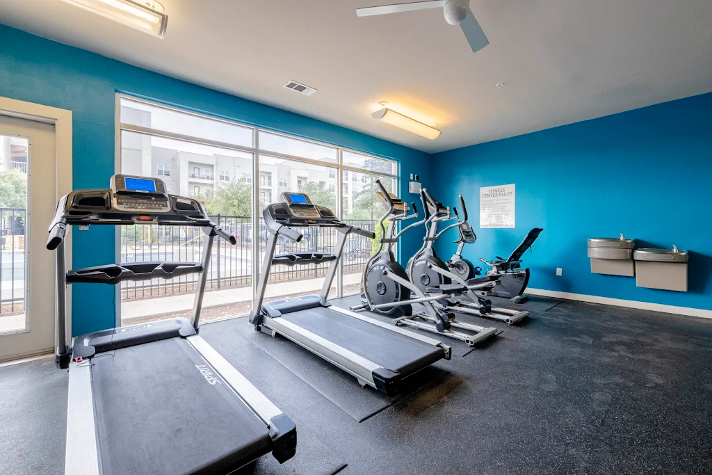 Community fitness center with treadmills, bikes, drinking fountains, and floor to ceiling windows for natural light