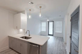 White stone counter island with sink & dishwasher and pendant lighting, behind which there is a livingroom with a patio door