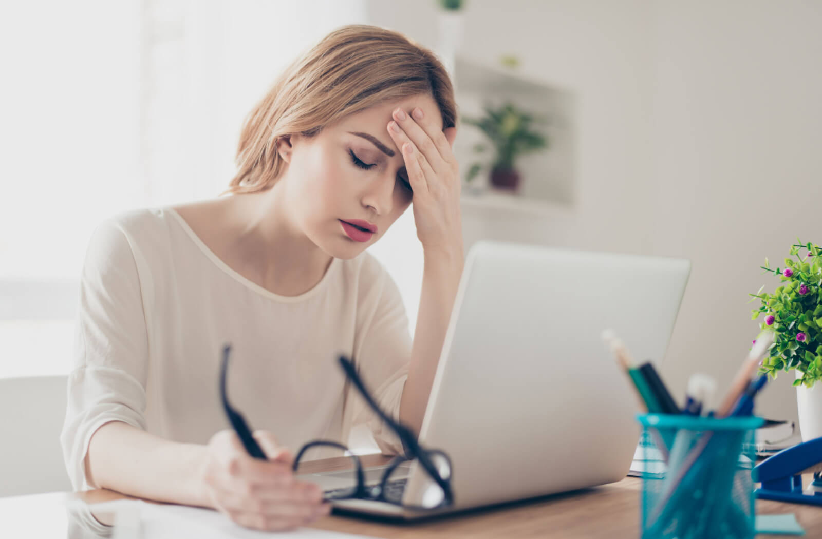 A lady in a white blouse is sitting in front of her computer while she is touching her forehead with her left hand and holding her eyeglasses in her right hand.