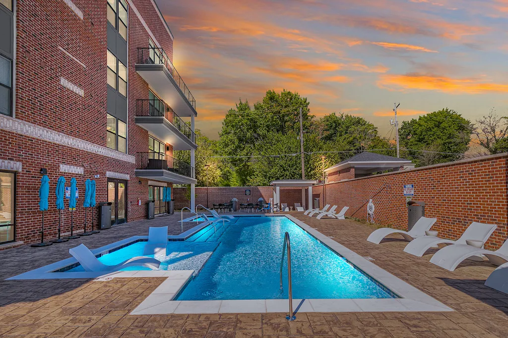 Swimming pool at dusk, next to apartment buildings, with sundeck featuring white lounge chairs surrounding