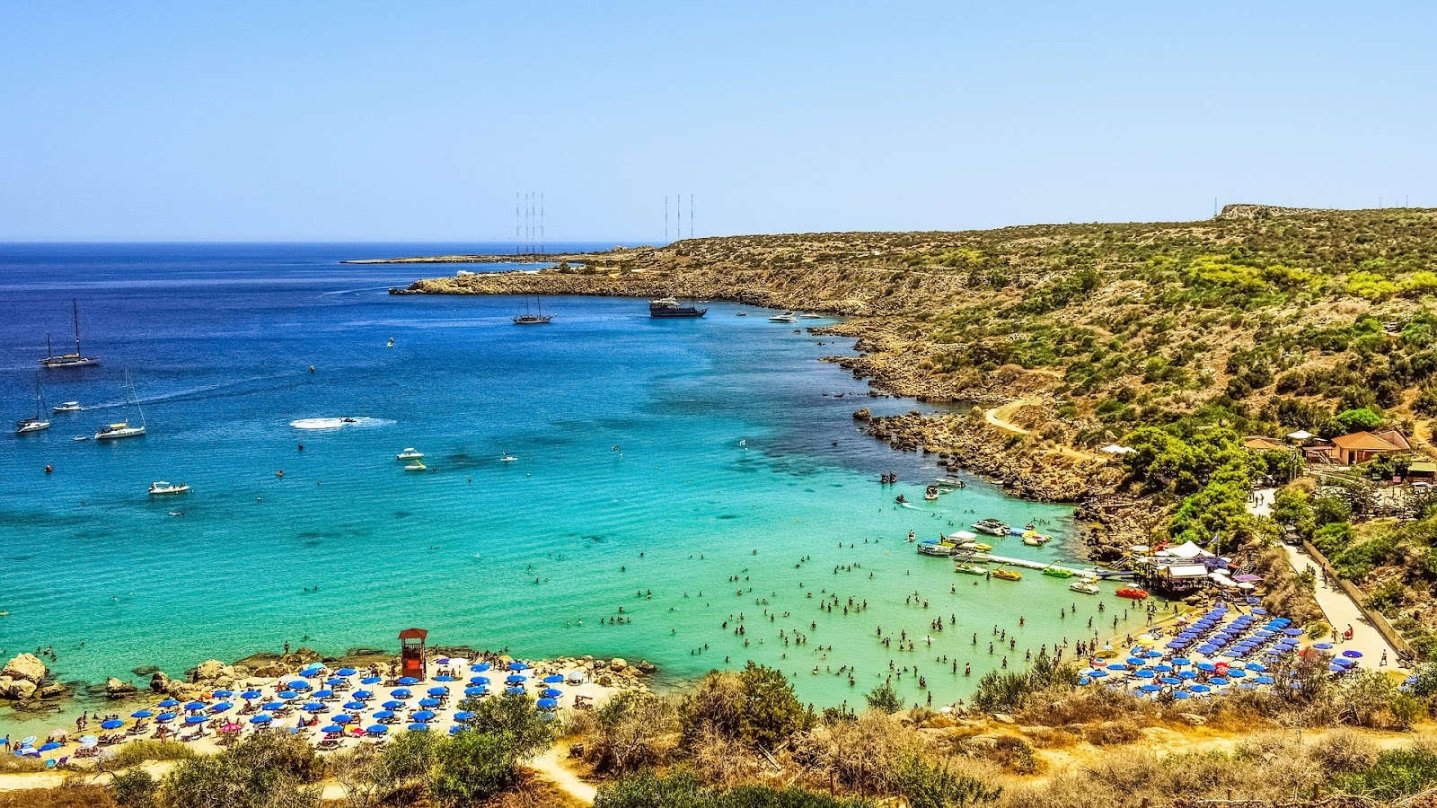 konnos small bay and two sandy beaches with blue sunbeds, tourists swimming in emerald green water. boats in distance and green trees around the shore