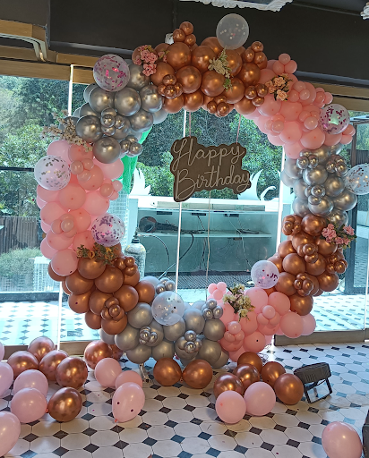 The image shows a birthday decoration setup featuring a large balloon arch in pink, rose gold, an… post image