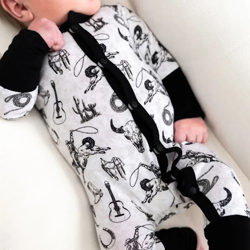 Close-up of a small reclining figure wearing a white snap-front footless onesie printed with western-themed illustrations on a pale cushioned background