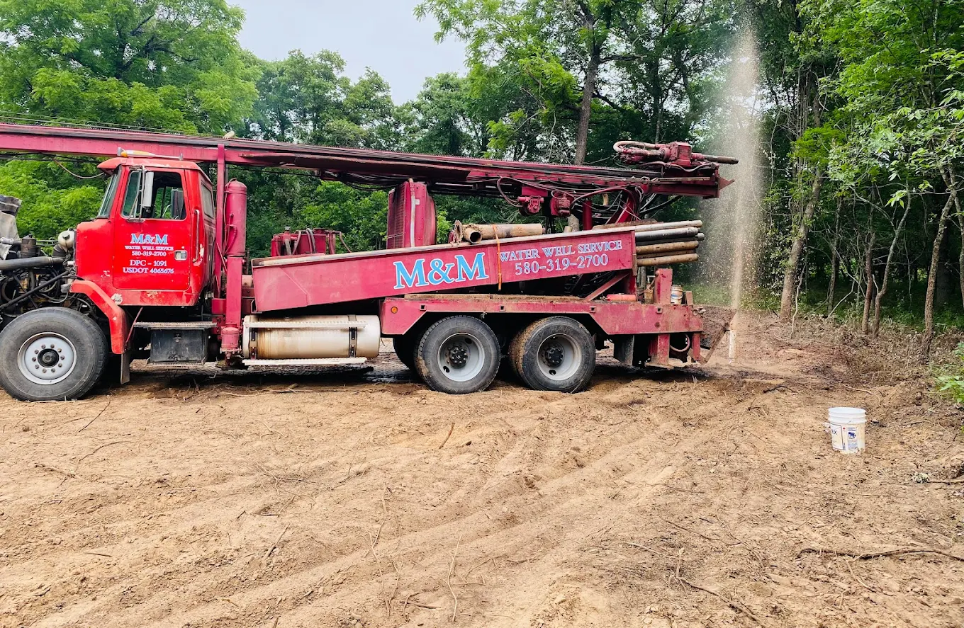 A red water well drilling truck is in operation, with visible mud and debris spraying from the drill site, showcasing ongoing well installation or maintenance.