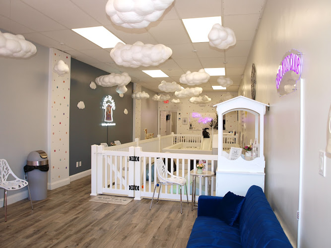 The indoor play area features whimsical decor with cloud-like structures hanging from the ceiling. The layout includes a play space enclosed by a white fence, creating a secure area for children. Seating is provided for adults, complemented by a plush blue sofa. Bright wall colors and soft flooring suggest a welcoming and safe environment for outdoor play. Within the fenced area, there are climbing structures and a ball pit noted as key elements for children's activities.