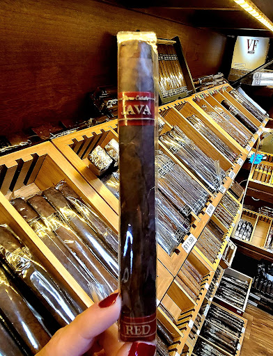 Hand holding a sealed cigar in cellophane in front of humidor shelves and organized trays