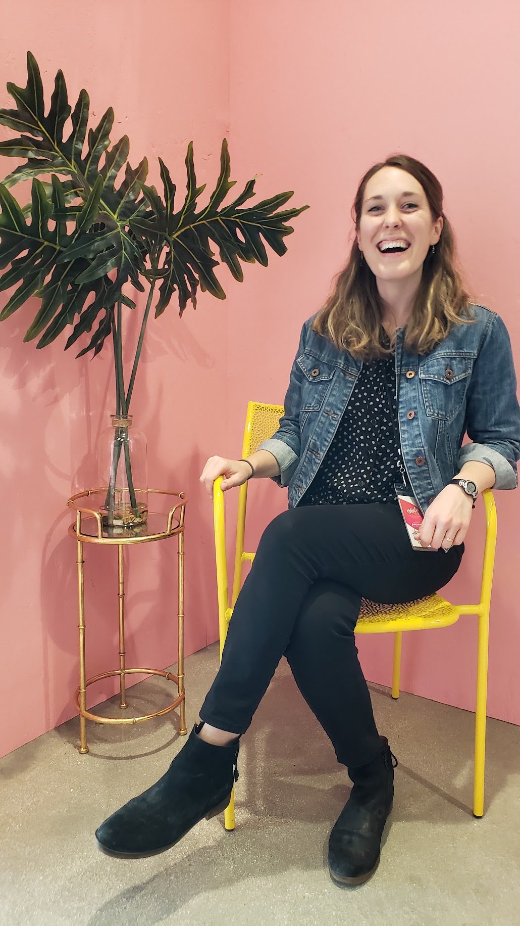 Women laughing while sitting in a yellow chair in front of pink walls and a large plant.