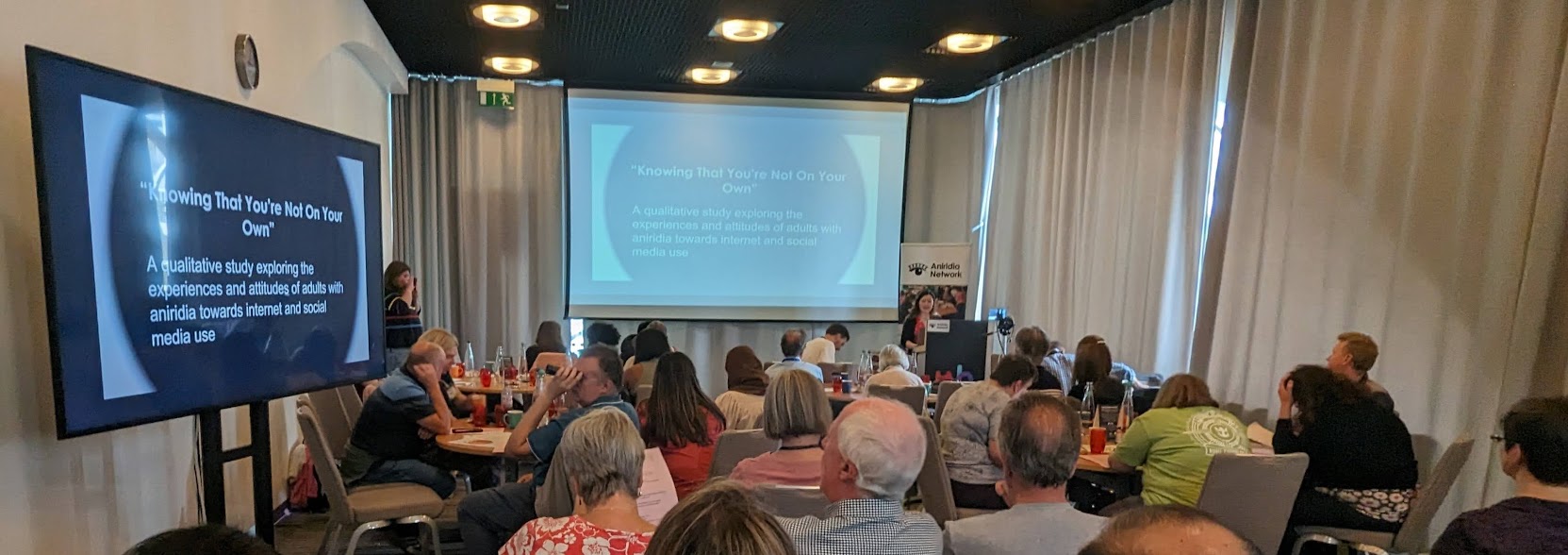 Conference session viewed from behind seated audience. Screens show s large black circle with white title text 'Knowing that you're not on wour own'