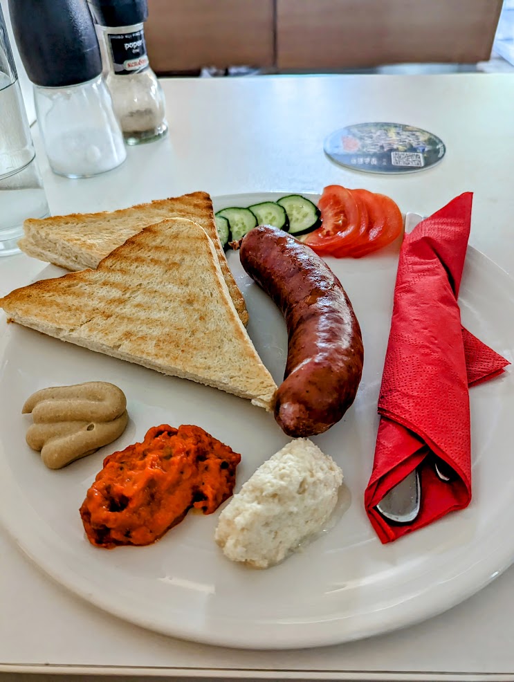 Sausage on white plate with two slices of bread and 3 sauces and some tomato and cucumber