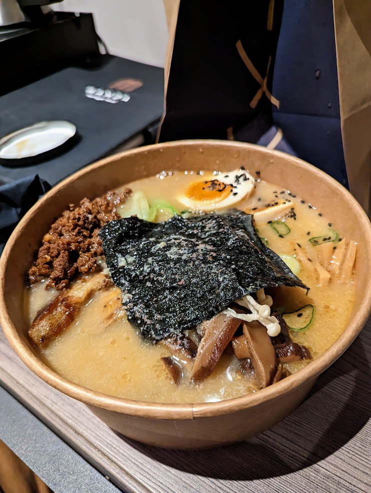 Bowl of cloudy ramen in cardboard container, seaweed slice on top