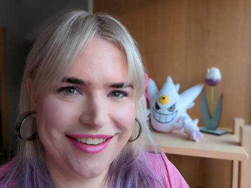 A woman smiling into the camera. She has blonde hair with the tips dyed purple, and is wearing pink eyeshadow and lipstick, large translucent hoop earrings, and a pink top. A plush toy of shiny Mega-Gengar looks on approvingly in the background.