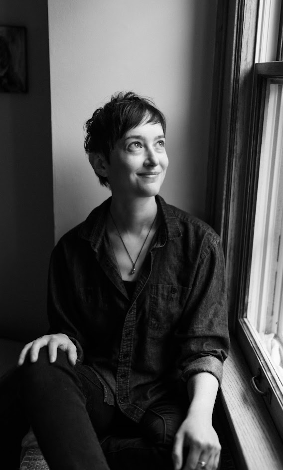 Black-and-white portrait of a person with short, dark hair and pale skin smiling and looking out a window