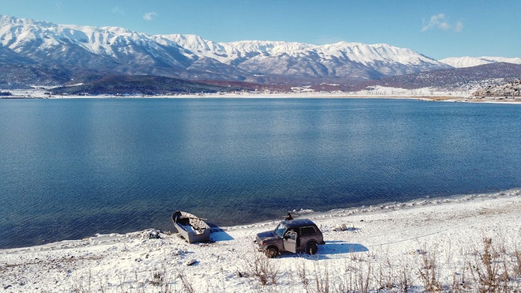 An aerial drone picture of a car and boat parked on the snow by big Prespa Lake West Shore, Korce County, Albania in the winter. Snowy mountains are in the distance