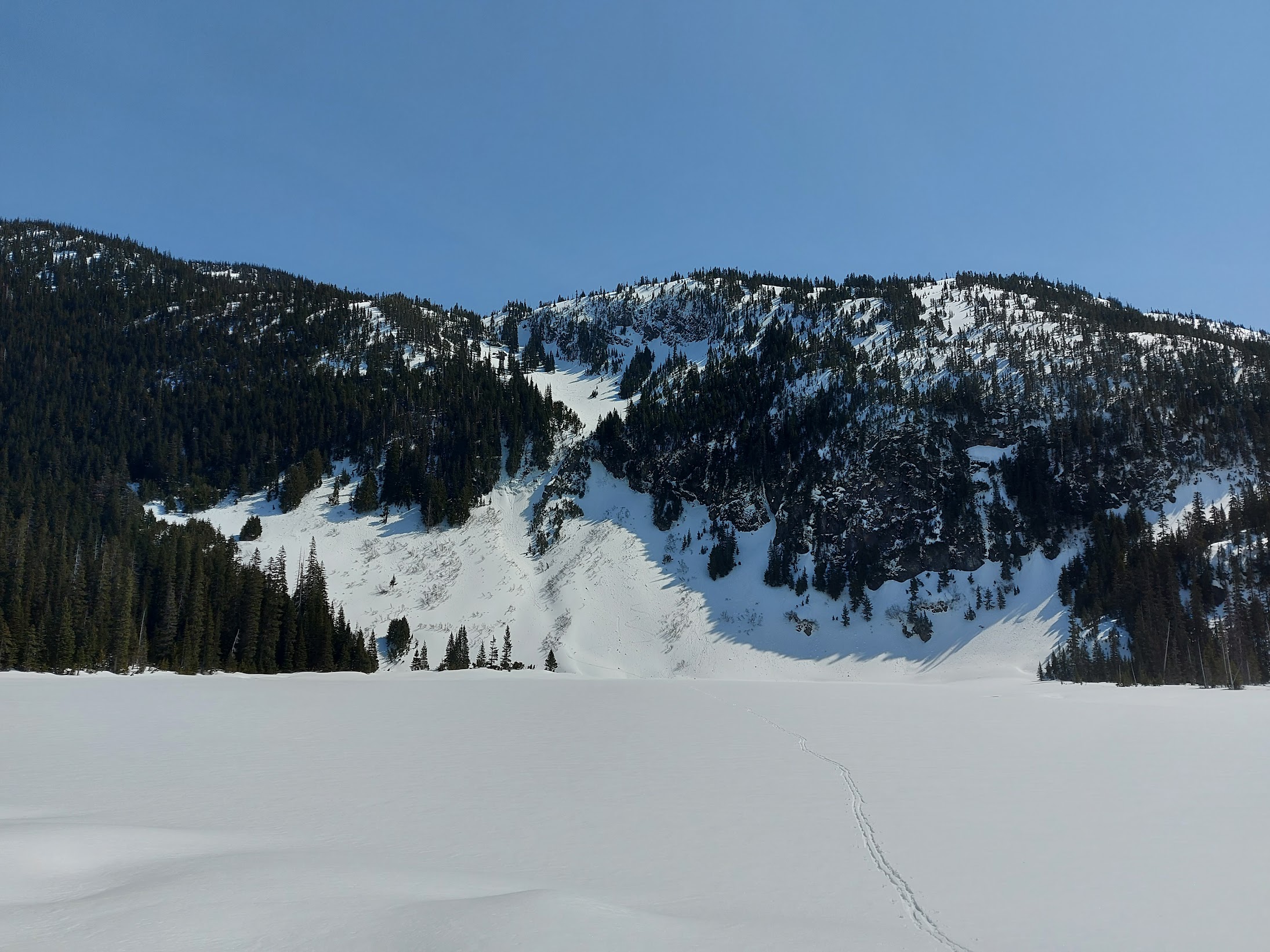 View across frozen lake with skin tracks. Cliff band with small snowy gully above lake.