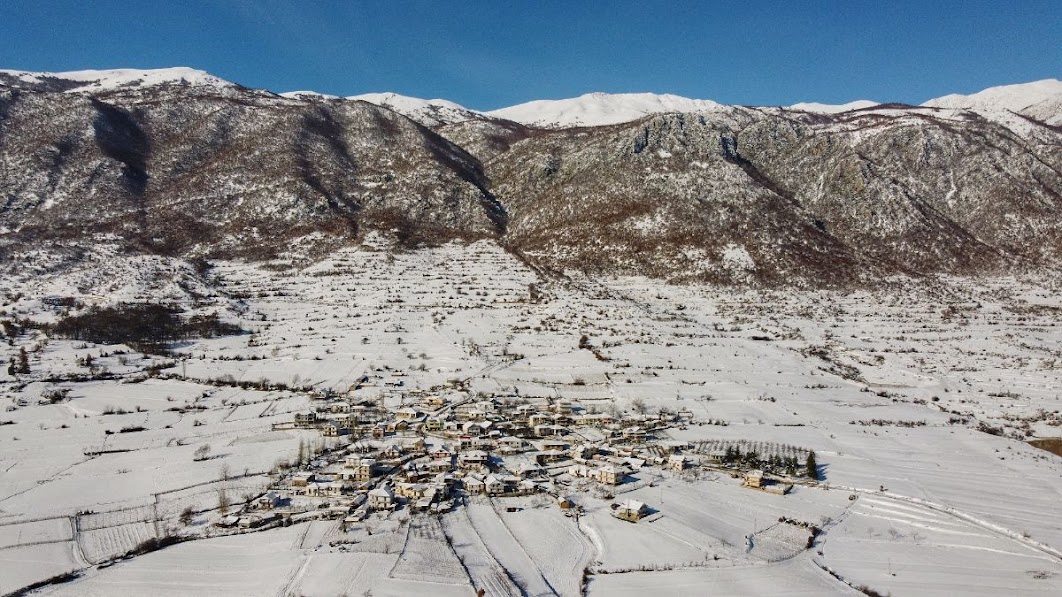 An aerial drone picture of a snow covered village near big Prespa Lake West Shore, Korce County, Albania in the winter. Snowy mountains are in the distance.