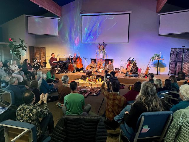Image is of a group of people sitting on chairs and on the floor while musicians sit on the stage playing music