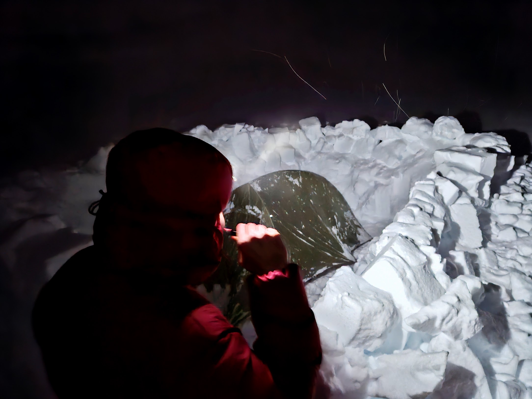 Skier brushing teeth in the dark, with a tent covered in snow, surrounded by a wind wall, in the background