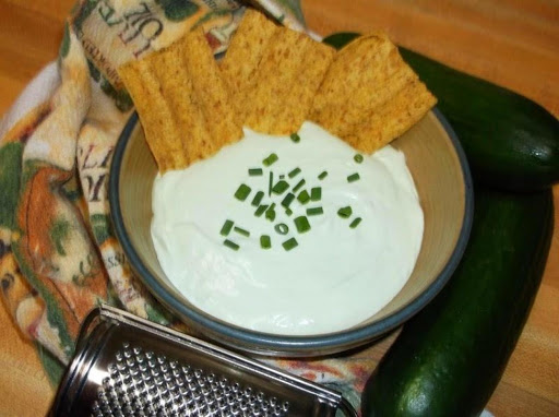 Bowl of benedictine spread with chips surrounded by cucumbers, grater and kitchen towel.