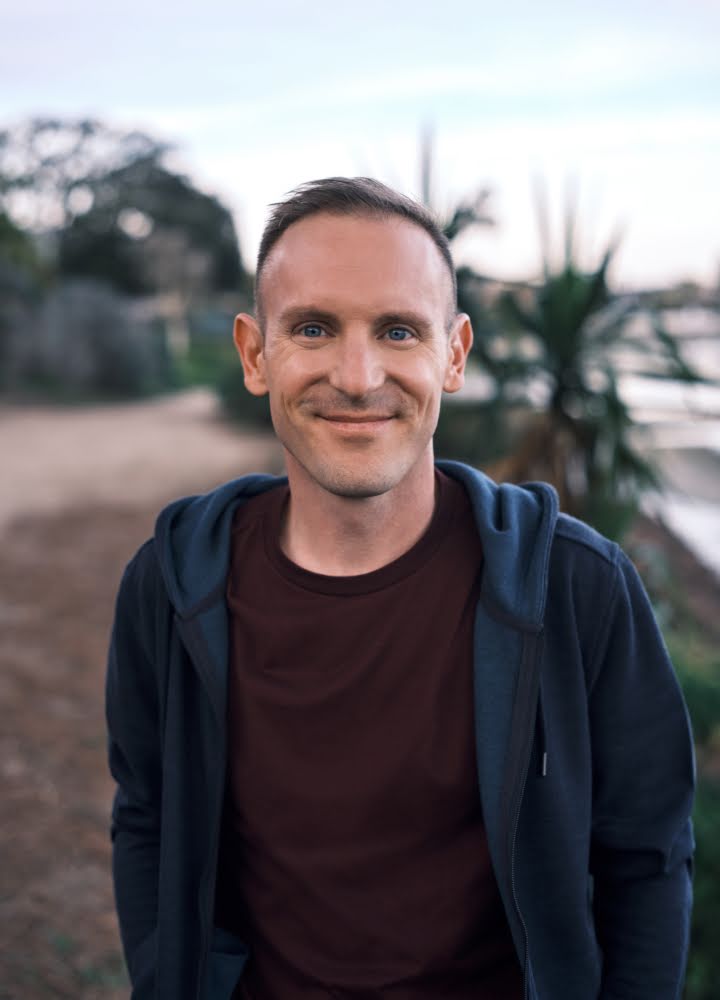 Portrait of John Callery in San Diego standing outdoors in front of trees and water with the background blurred slightly