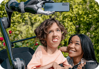 A white person with wavy brown hair sits in their motorized wheelchair with a robot arm attachment holding an Android phone. A Black woman crouches next to them and smiles up at the Android phone.