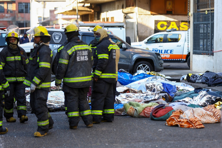 Firefighters stand next to covered bodies of victims at the scene of a deadly blaze in Johannesburg on August 31 2023.